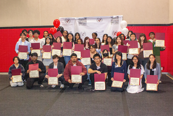 Group of smiling high school seniors of many races and ethnicities stand in rows holding up scholarship certifications.