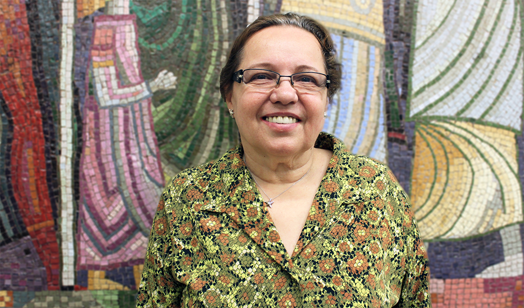 Woman in green and brown shirt stands in front of a colorful mosaic wall.