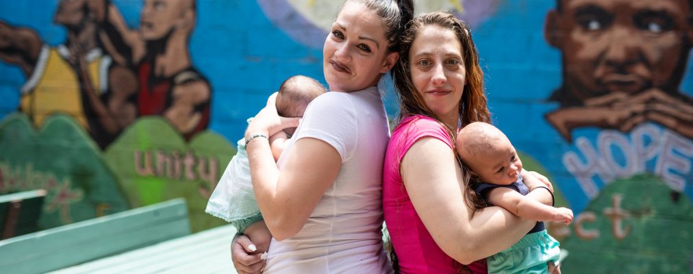 Two mothers stand back to back holding babies