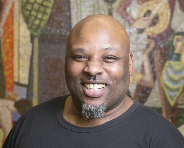 Korey Smith, a Black man with bald head and trimmed beard, smiles for a portrait in the Community Consultation Center community room
