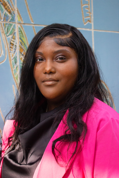 Breahna Watkins-Cole, a Black woman, wears a bright pink blazer and her hair down in long curls.
