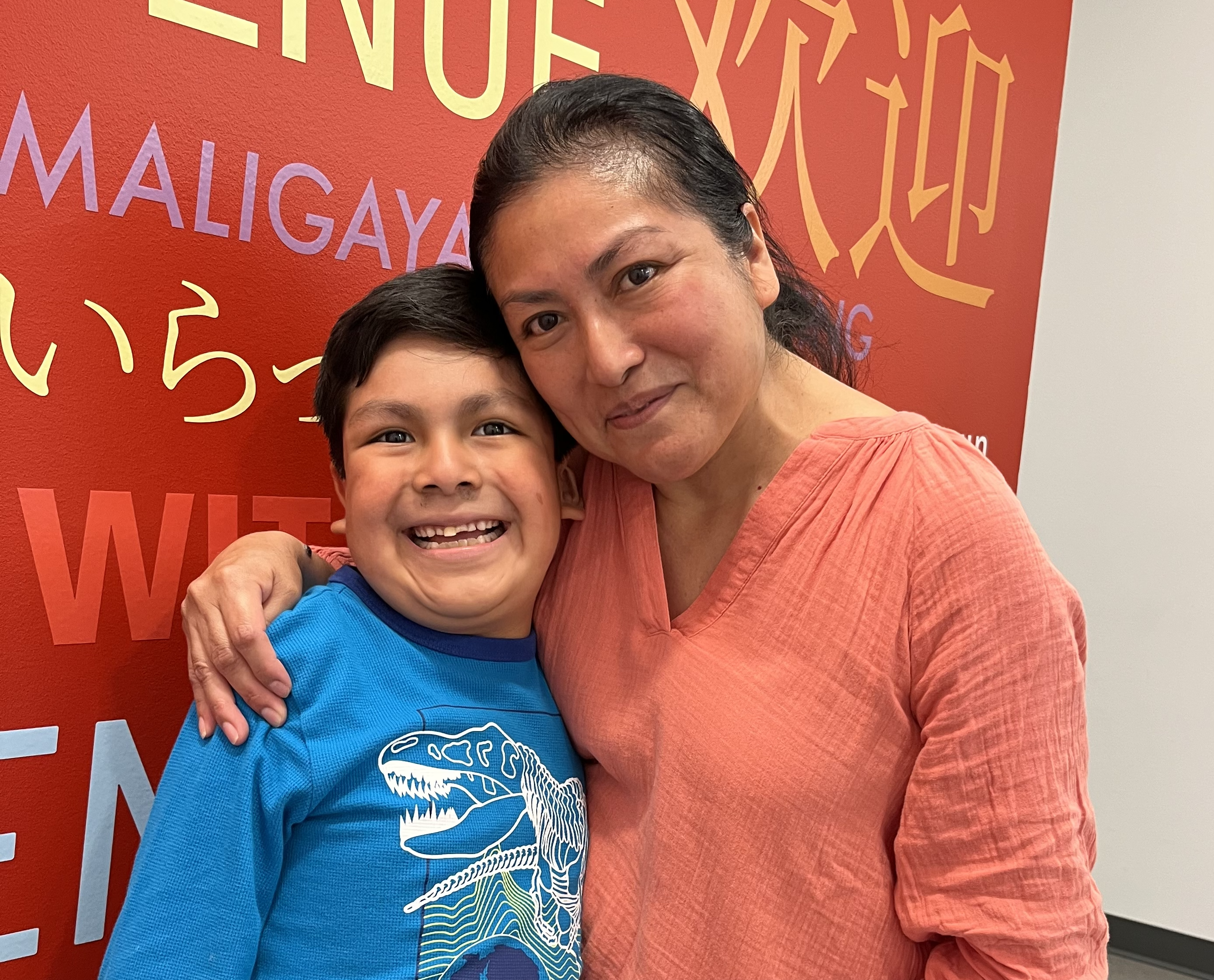 A mother holds her arm around her son in front of a colorful mural that says, "Welcome," in different languages.