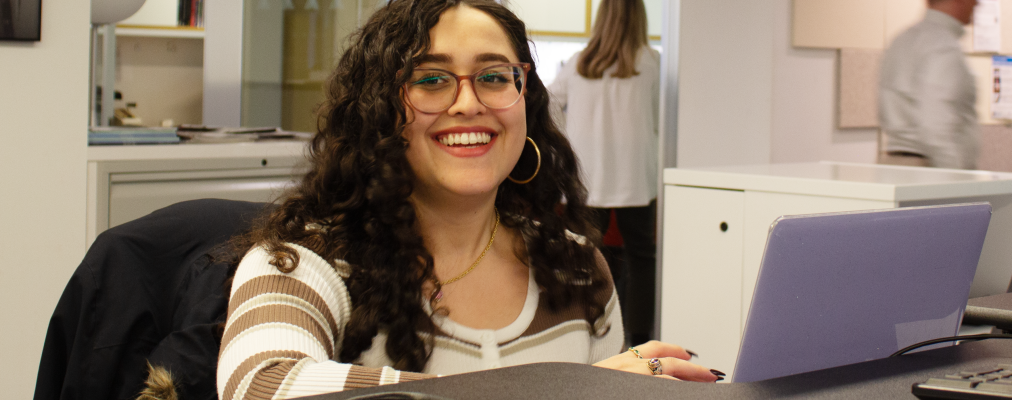 A woman smiling into the camera white seated in front of their laptop