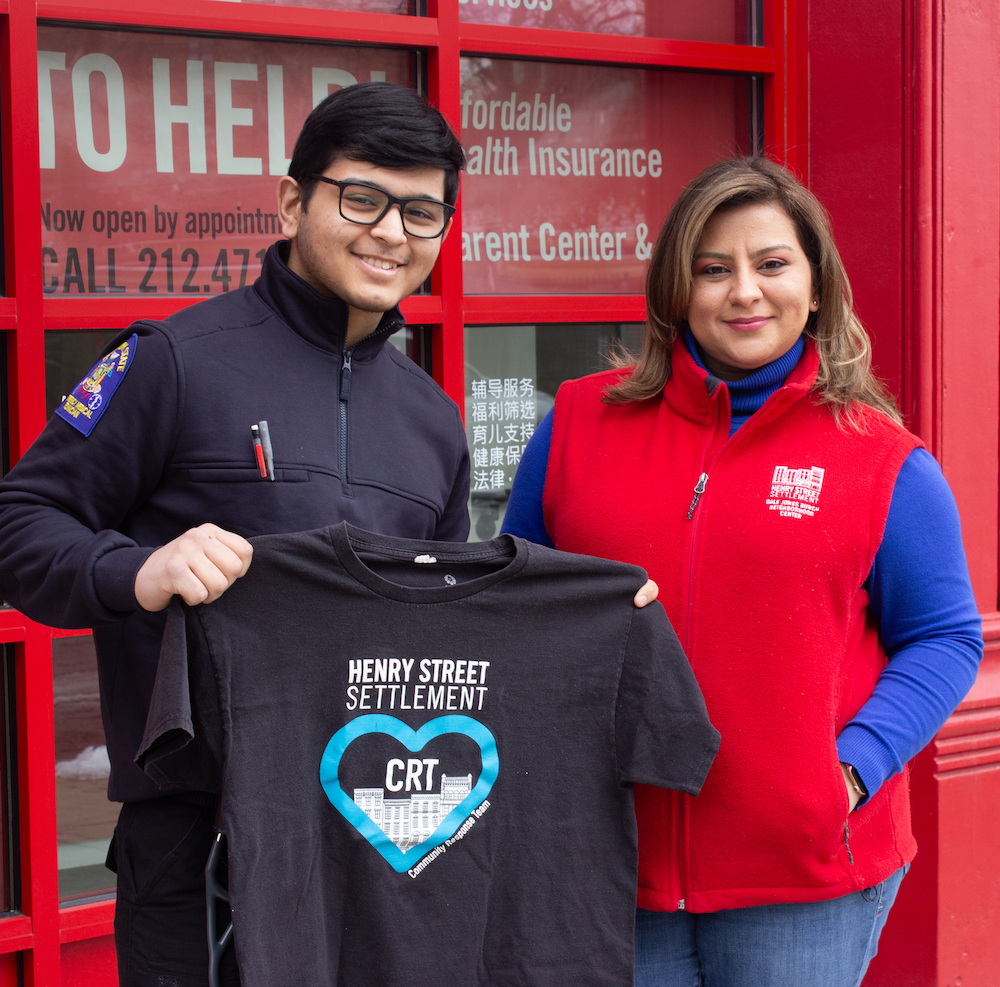 A young man in an EMT uniform holds up a t-shirt that says, "Henry Street Community Response Team." His mother stands at her side in a red vest with the Henry Street logo.