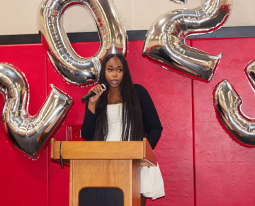 A young Black woman with long braided hair speaks into a microphone behind a podium. Big silver balloons that spell out 