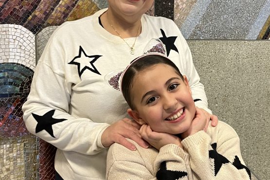 A mother stands with her hands on her daughter's shoulders. They both smile and wear matching cream sweaters with black stars embroidered.