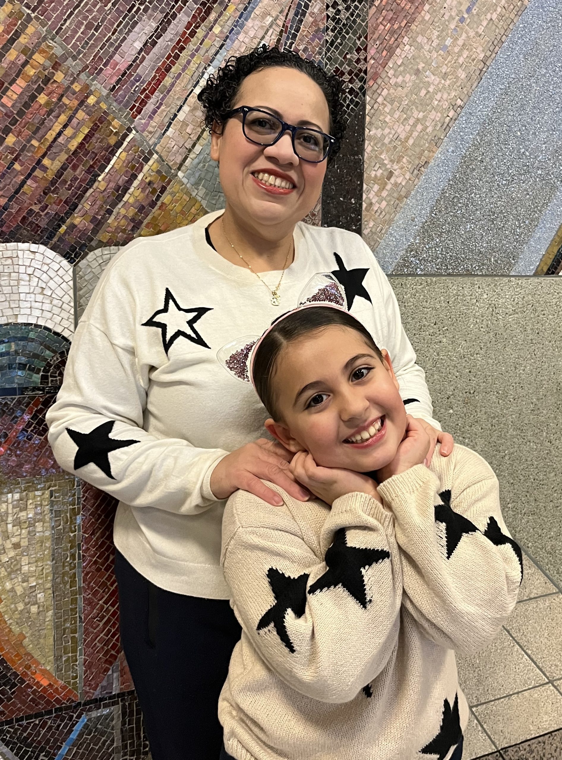 A mother stands with her hands on her daughter's shoulders. They both smile and wear matching cream sweaters with black stars embroidered.